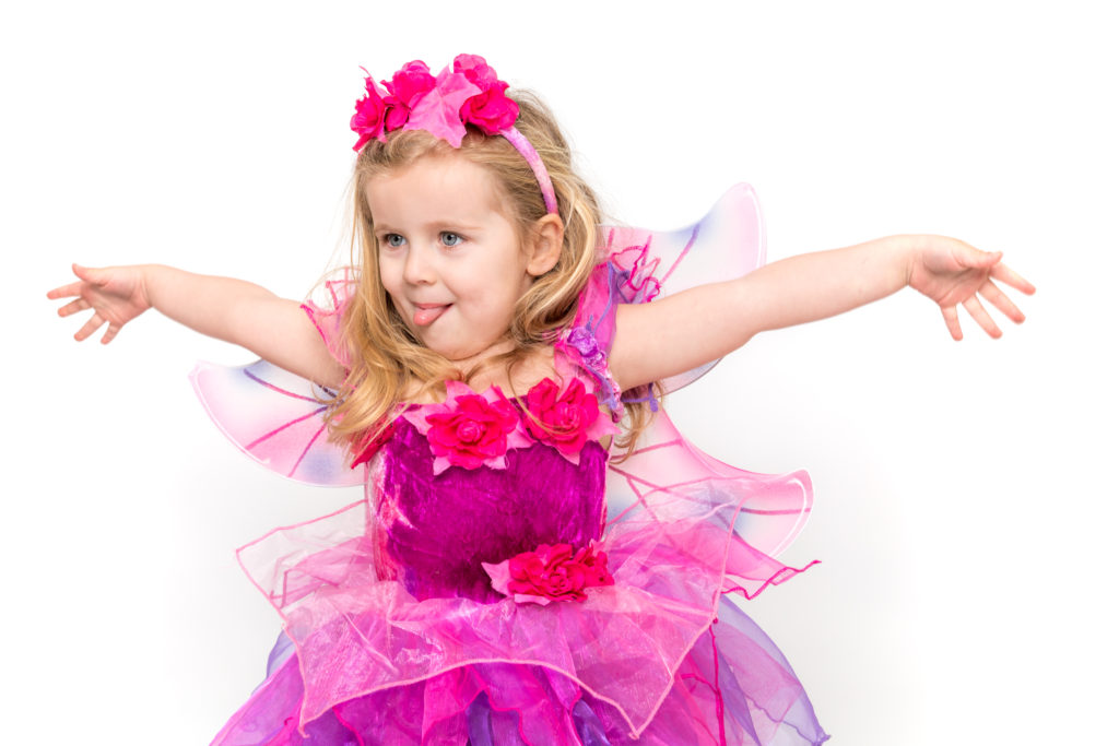 A small girl dressed as a fairy opens her arms wide and sticks out her tongue. Studio photo shoot.